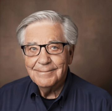 Elderly man with silver hair and glasses smiles warmly against a neutral background, conveying a friendly and approachable demeanor.