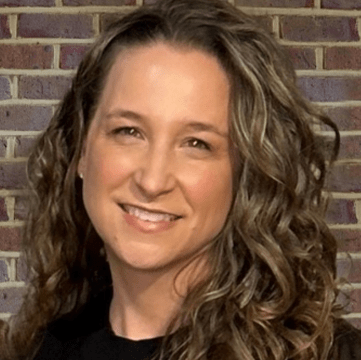 Woman with curly hair smiling, set against a brick wall background. This image highlights a friendly, approachable demeanor.